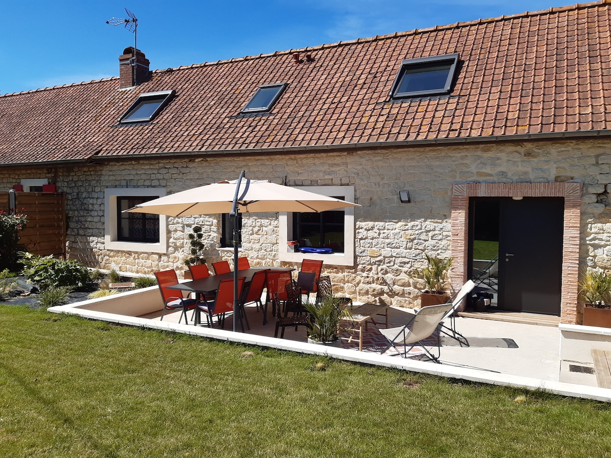 An outdoor seating area is displayed with a large umbrella providing shade over a table surrounded by several chairs. The house features a stone facade and multiple skylights. A well-maintained lawn and a sun lounger complete the inviting outdoor space.