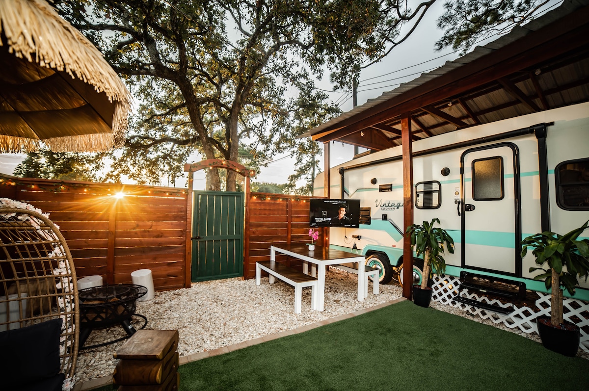 An outdoor space is showcased featuring a quaint RV parked next to a cozy fire pit area. A long table with benches sits on a grassy surface, surrounded by wooden fences and tropical plants. Evening sunlight filters through trees, creating a warm ambiance.