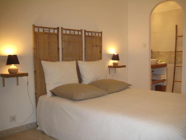 A light-filled bedroom features a spacious bed with plump pillows and bedside tables with lamps. Soft tones in the décor create a soothing ambiance. An archway leads to an adjoining bathroom, with a glimpse of natural materials evident in the design.