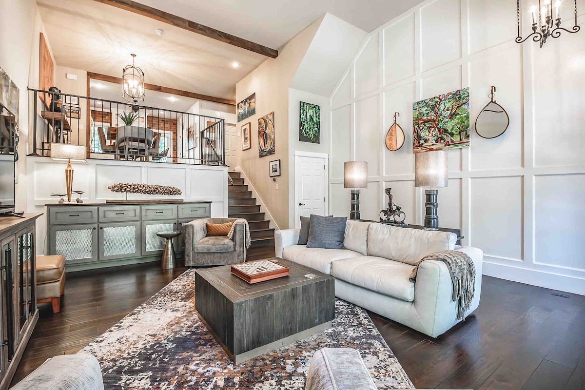 A comfortable living area is highlighted by a spacious white sofa, a coffee table, and a decorative area rug. Artwork adorns the walls, and natural light floods the space from large windows. An inviting staircase leads to the upper level, enhancing the open feel.