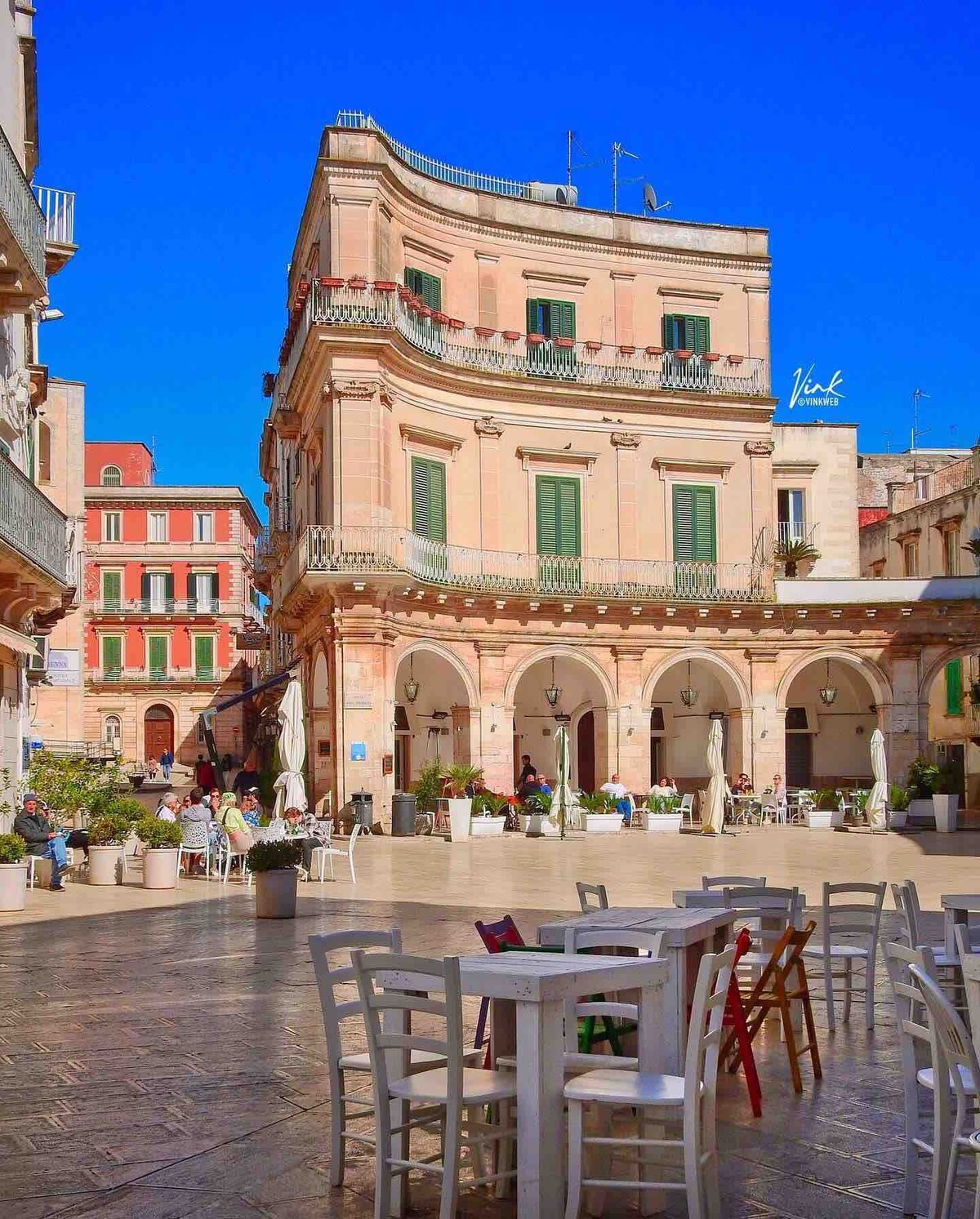 A vibrant square is characterized by a grand, historic building featuring green shutters and a mix of architectural styles. White tables and chairs are arranged beneath the colonnades, with potted plants adding a touch of greenery. The bright blue sky enhances the lively atmosphere.