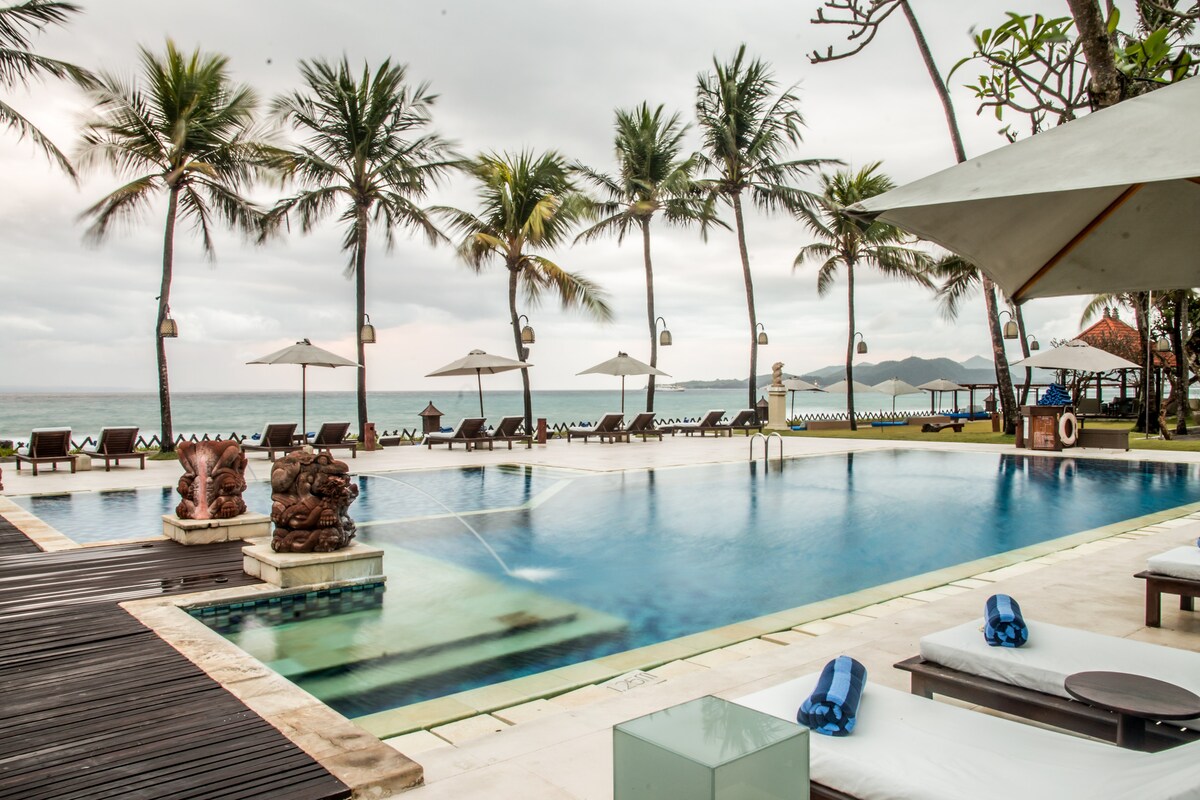An outdoor swimming pool is surrounded by palm trees, with several sun loungers positioned on a wooden deck. Umbrellas provide shade over the lounge areas, and a gentle view of the ocean can be seen in the background.