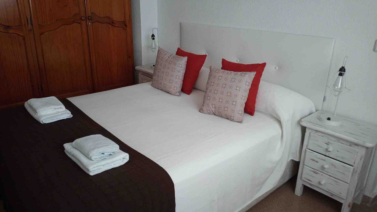 A spacious bed is dressed in crisp white linens, complemented by decorative pillows in red and patterned fabric. Two small bedside tables with lamps are positioned on either side, while a wooden wardrobe is visible in the backdrop.