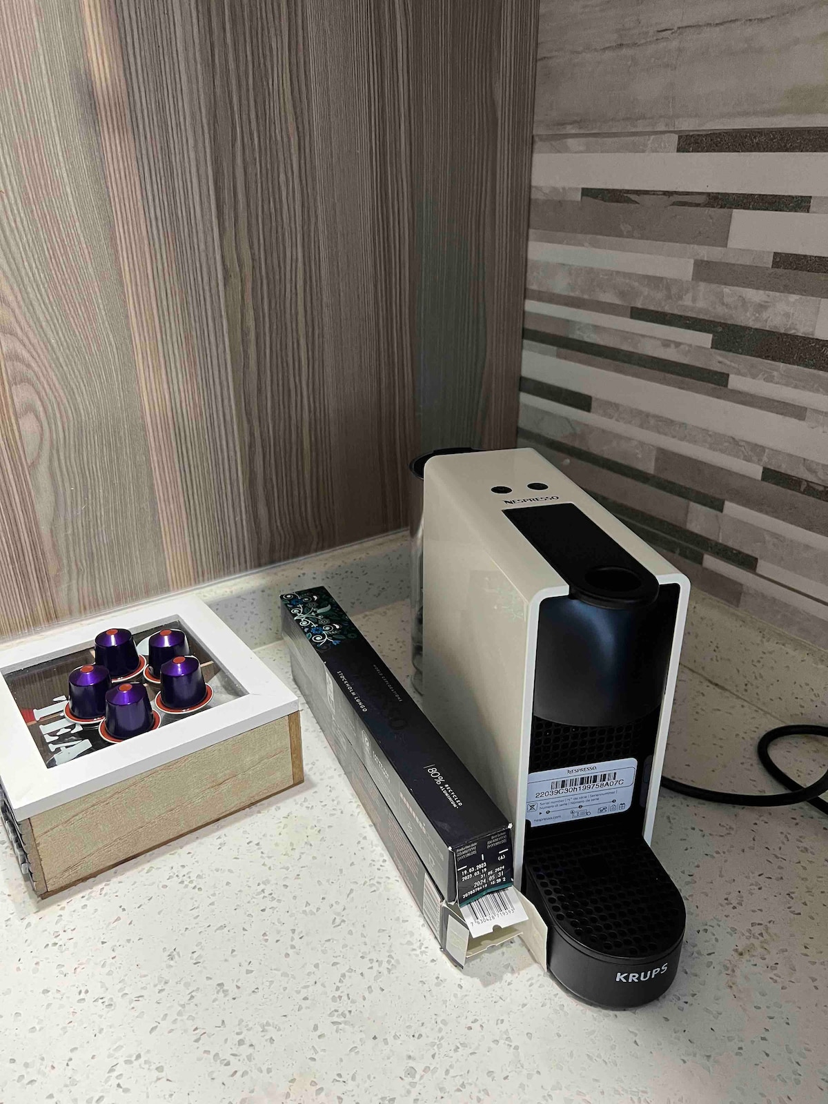 A sleek coffee machine is displayed next to a decorative box containing several coffee pods. The countertop is complemented by a stylish backsplash featuring neutral tones and subtle patterns, enhancing the modern aesthetic.