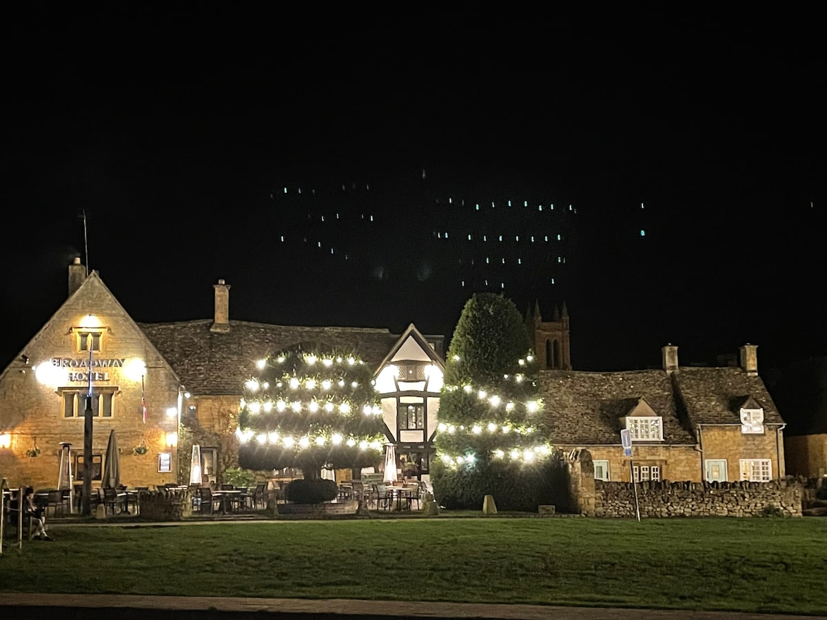 A charming building is illuminated at night, showcasing its warm stone façade and large outdoor seating area. String lights adorn a tree, enhancing the welcoming ambiance. In the background, the silhouette of a church tower adds to the historic character of the setting.