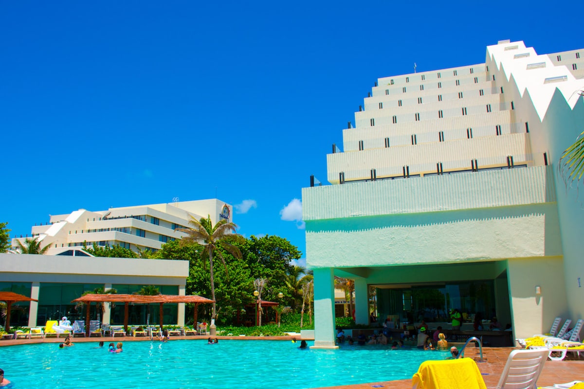 A large pool area is showcased, surrounded by palm trees and shaded lounge chairs. The building in the background features a unique architectural design with multiple levels and large windows. Clear blue skies enhance the vibrant atmosphere of the resort.