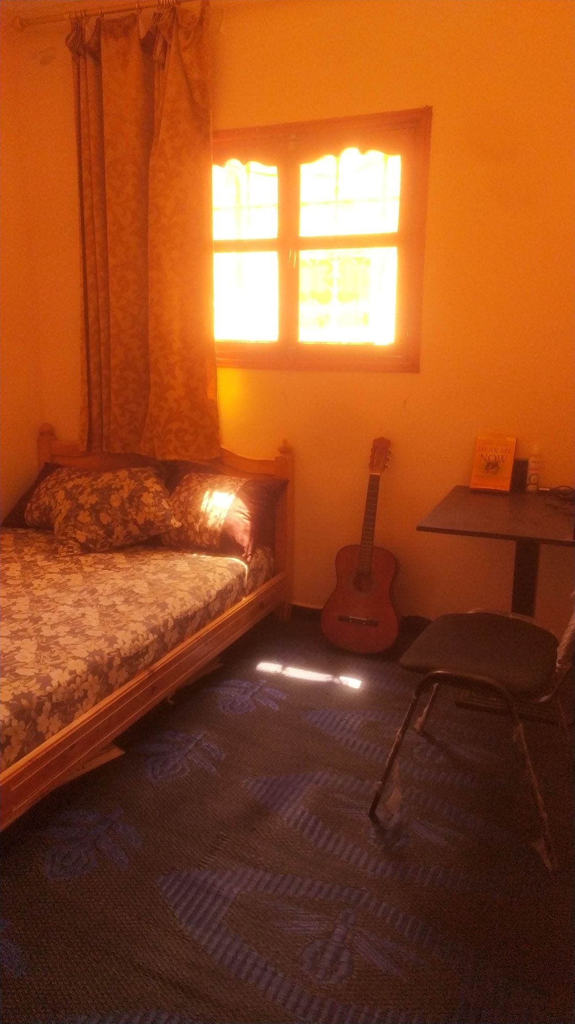 A cozy bedroom features a single bed adorned with patterned bedding. Natural light streams through a window with warm orange curtains, illuminating the room. A small desk is positioned nearby, along with a guitar resting against the wall.