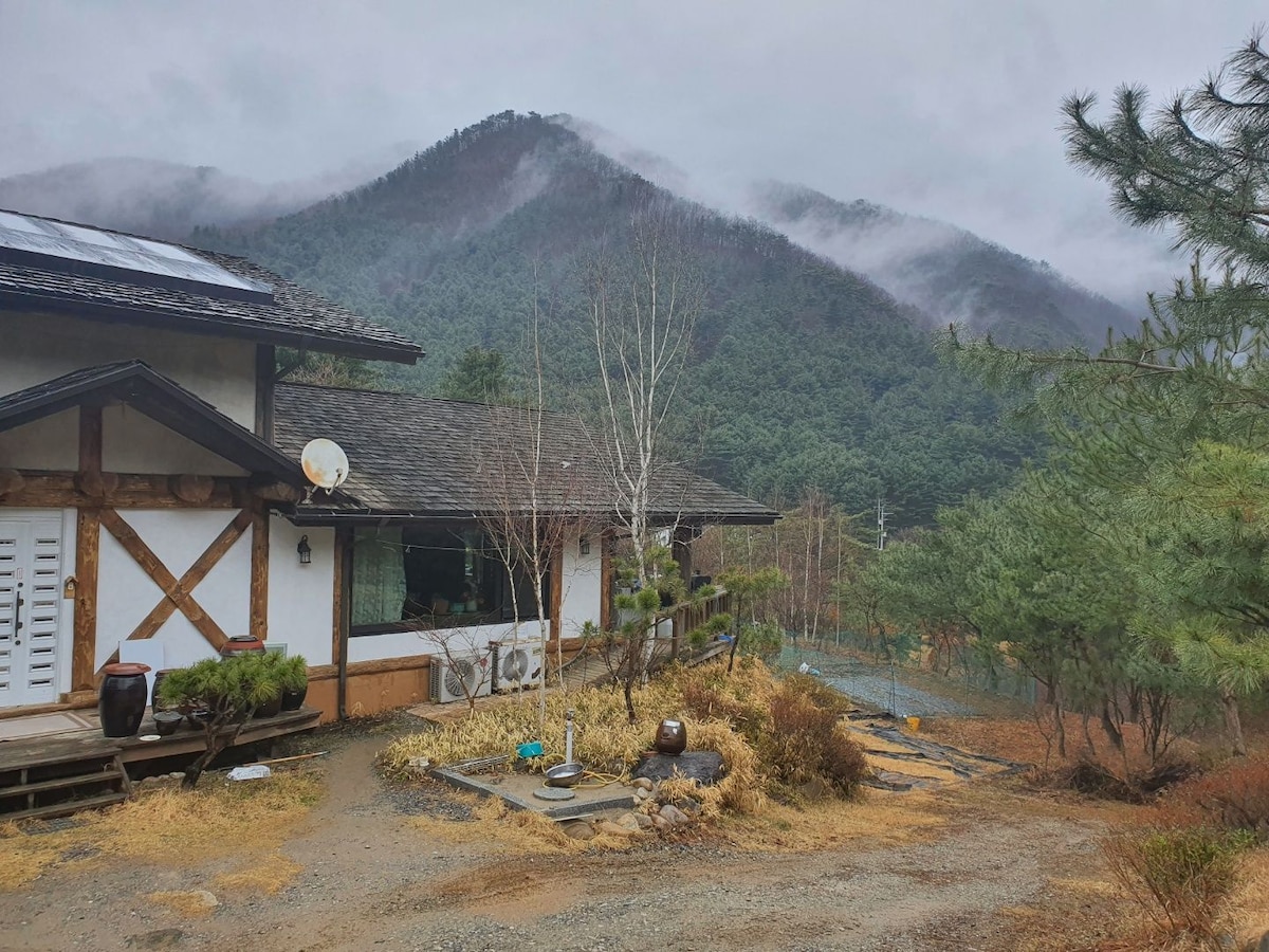 A serene exterior view showcases a rustic house set against a backdrop of misty mountains. The well-maintained yard features a stone pathway leading to the entrance, with scattered dry foliage and evergreen trees adding to the natural setting.