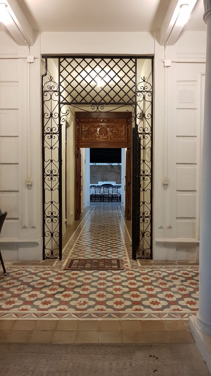 A hallway features intricate tile patterns and high ceilings, leading to an open space with dining seating visible in the background. Ornate wrought iron gates separate the hallway from the inviting area, while soft lighting enhances the architectural details.