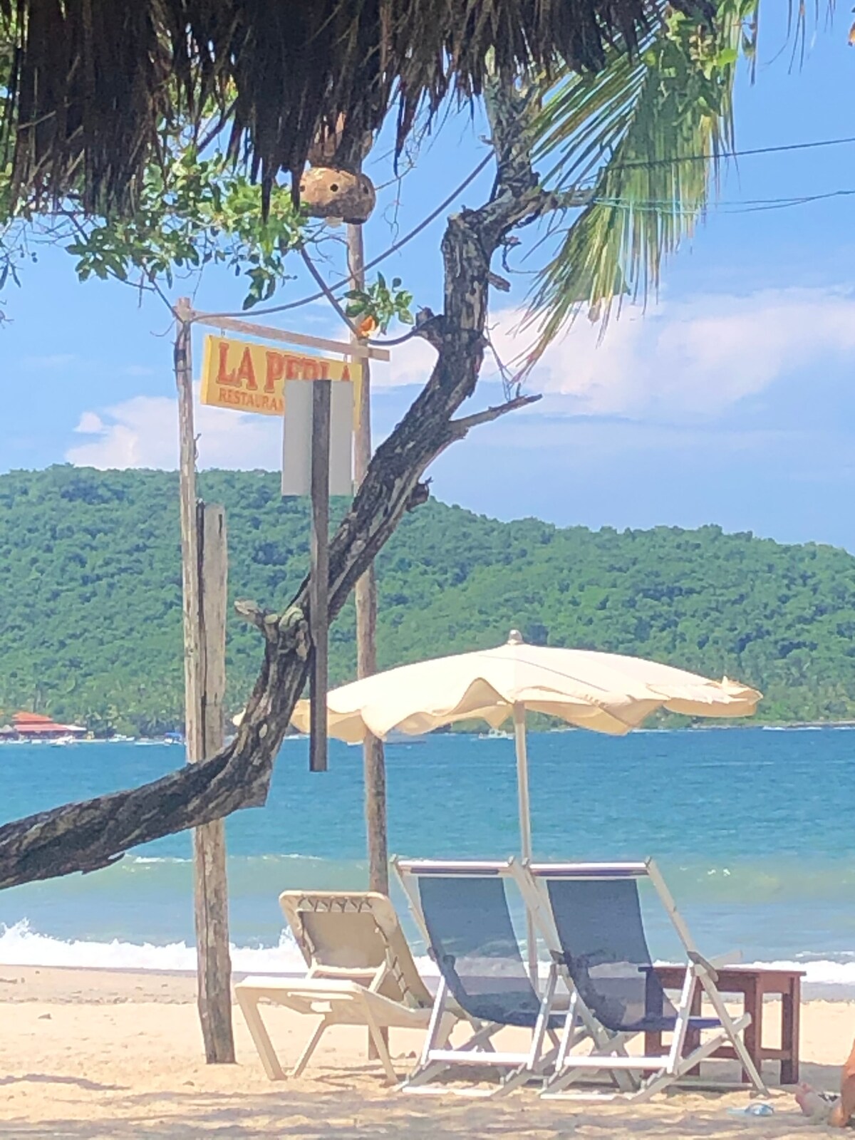 A tranquil beach scene is captured, featuring a white umbrella providing shade over two lounge chairs. A rustic sign reads 'La Perla' in the background, with lush green hills rising in the distance. The turquoise water gently meets the sandy shore under a clear blue sky.