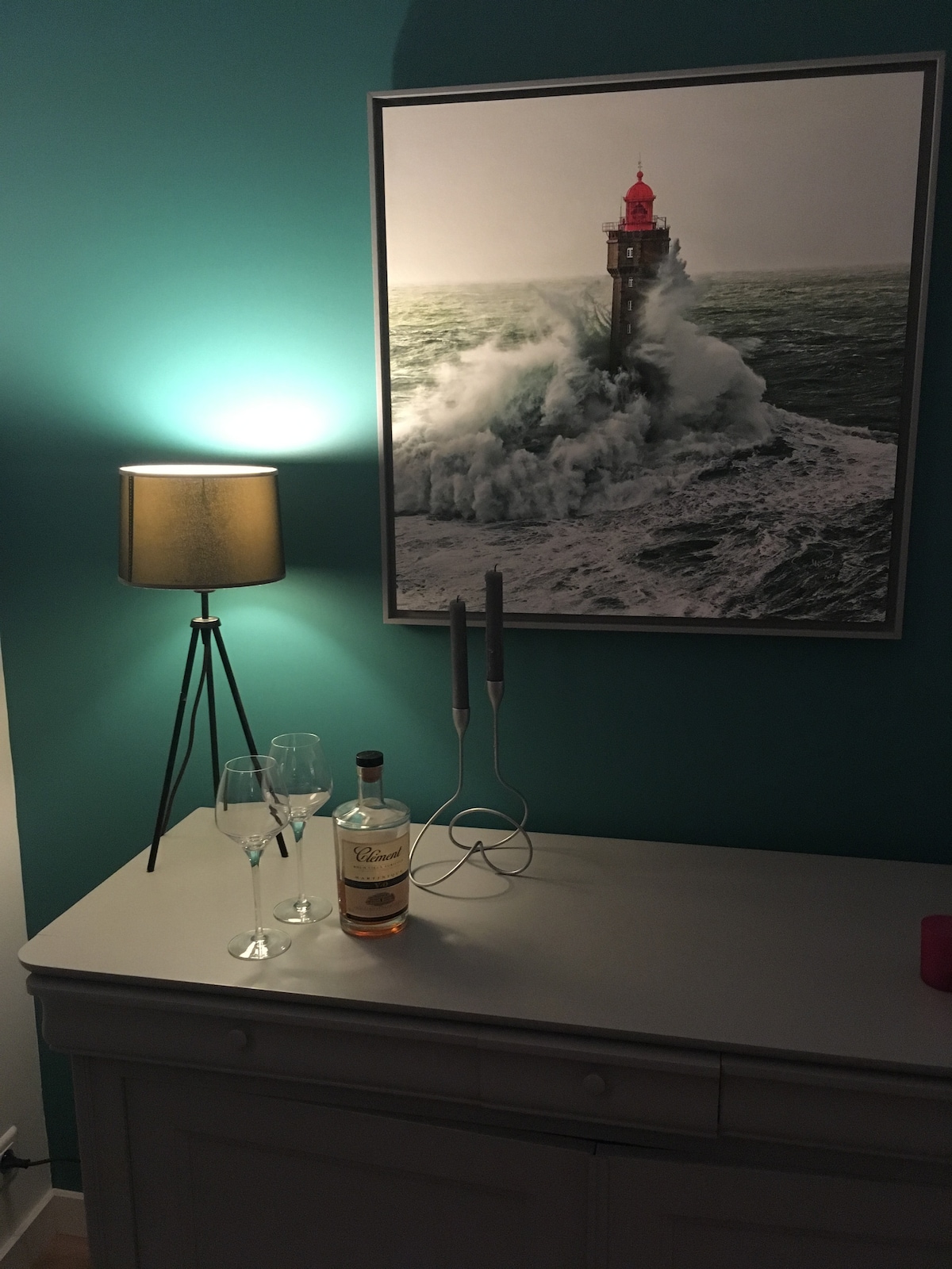 A modern lamp emits soft light beside a decorative piece featuring a lighthouse on a turbulent sea. A bottle of wine is placed on the surface, accompanied by two wine glasses and a candle holder with two candles, all set against a bold turquoise wall.