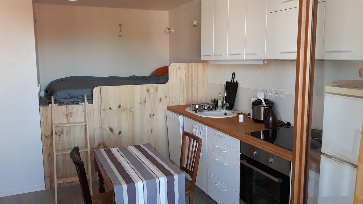 A functional kitchen area features a wooden countertop with a variety of kitchen utensils and appliances. A dining table with striped tablecloth complements the space. Above, a sleeping loft with a neatly made bed is partially visible, separated by a wooden partition.