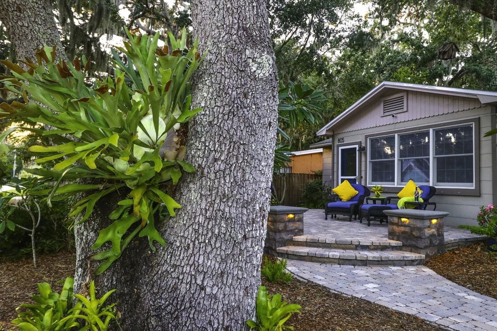 A charming bungalow is framed by lush greenery and mature oak trees. A paver deck features seating with yellow cushions, inviting relaxation. Decorative lanterns provide soft lighting along the pathway, enhancing the outdoor space's natural beauty.