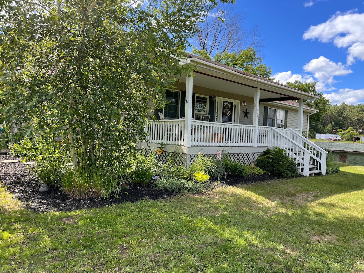 A welcoming front porch is visible, featuring a white railing and steps descending to an expansive lawn. Surrounding flowering plants and shrubs add greenery, while a star decoration is placed on the house's façade. A blue sky with fluffy clouds provides a serene backdrop.