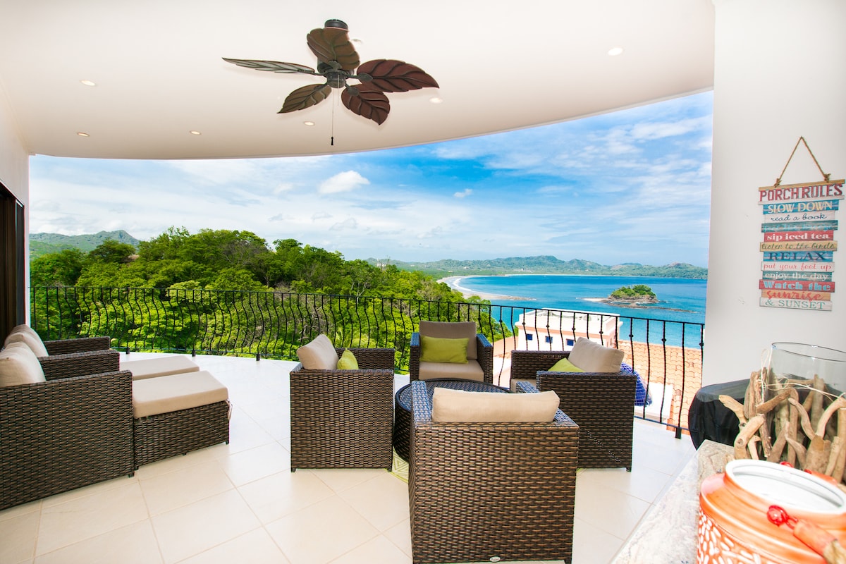 A spacious terrace showcases comfortable seating arranged to provide optimal views of the surrounding landscape, including the Pacific Ocean and lush greenery. Natural light floods the area, and a ceiling fan offers airflow, enhancing relaxation during warm days.