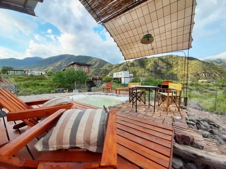An outdoor seating area features wooden lounge chairs and a table set beneath a fabric canopy. The surrounding landscape includes rolling green hills and mountains, providing a serene backdrop. A hot tub is visible nearby, adding to the relaxed atmosphere of the space.