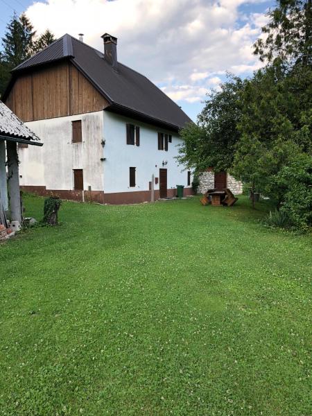 A two-story stone house with a wooden accent roof is surrounded by a lush green lawn. A shaded outdoor dining area, featuring a table and wooden benches, is positioned on the grass. Trees and shrubs frame the yard, creating a serene outdoor space.