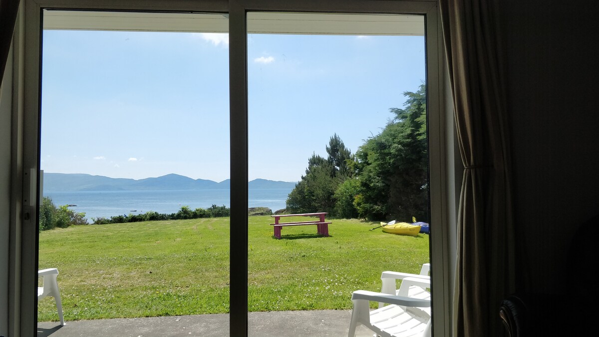 A view from the cottage's sliding glass door showcases a lush green lawn leading to Kenmare Bay. A pink picnic table is positioned on the grass, surrounded by trees and hills in the distance. Sunlight illuminates the scene under a clear blue sky.