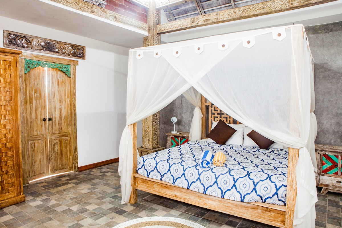 A spacious bedroom features a wooden four-poster bed draped with light fabric. A patterned blue and white bedspread adds a pop of color. Natural stone flooring complements the rustic wooden wardrobe and decorative elements on the walls, enhancing the room's tranquil ambiance.