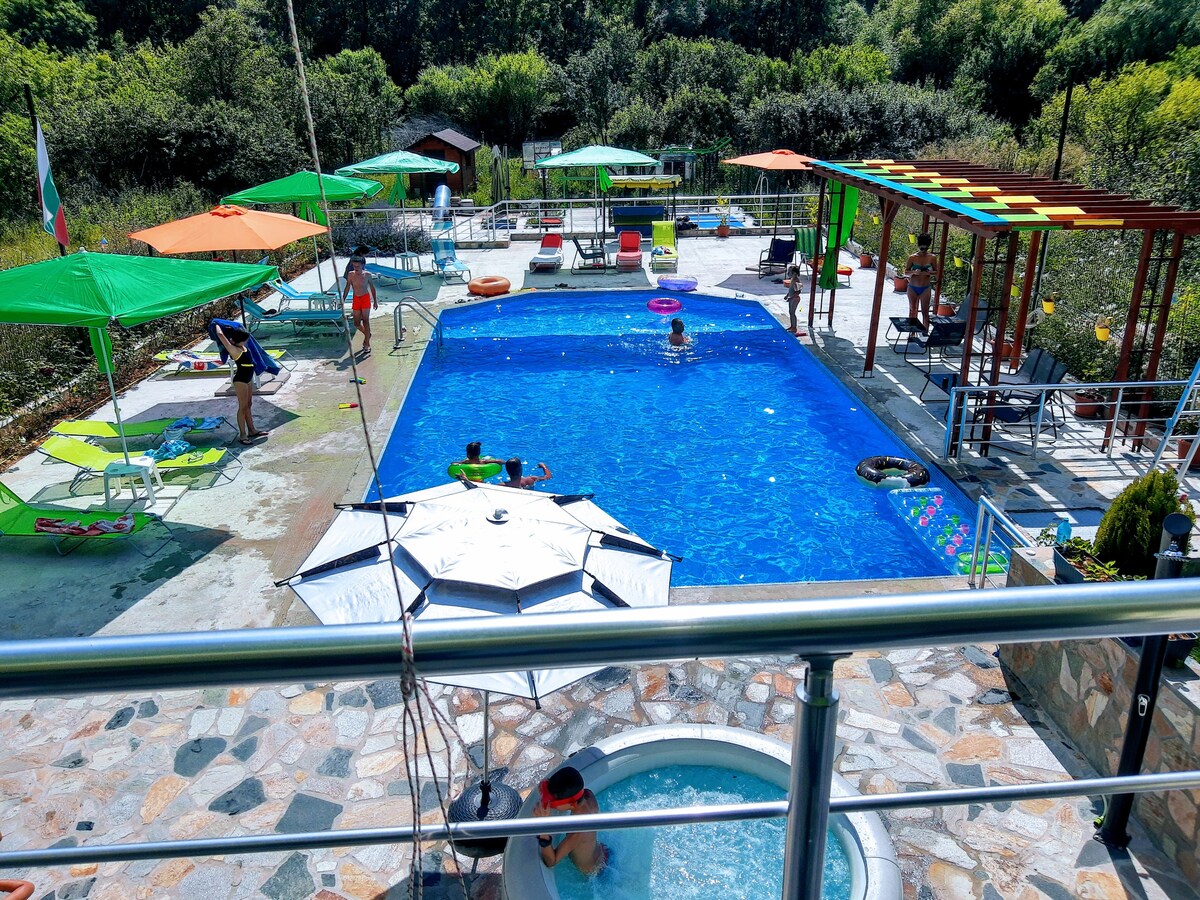 A resort area is shown, featuring a large swimming pool surrounded by lounge chairs and colorful umbrellas. A hot tub is placed to one side, with greenery visible in the background. People are observed enjoying various activities in and around the water.