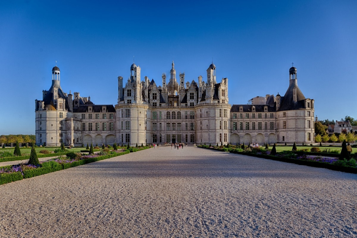 The stunning architecture of Château de Chambord is displayed across a grand façade, featuring intricate towers and ornate details. A vast gravel courtyard leads up to the entrance, while manicured gardens with vibrant landscaped borders enhance the setting under a clear blue sky.