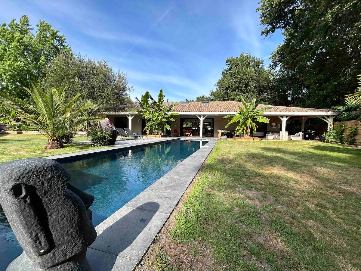 A spacious villa is showcased with a large swimming pool lined by tropical plants. The covered terrace extends from the villa, offering shaded seating areas. Surrounding greenery and a well-maintained lawn provide a tranquil outdoor space, enhancing the villa's serene atmosphere.