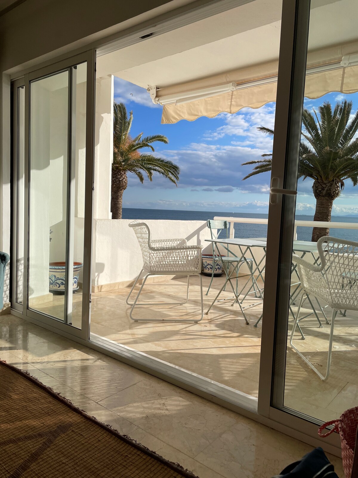 A glass door provides a view of a spacious terrace featuring outdoor seating. The terrace is bordered by palm trees and overlooks the sea, offering glimpses of blue sky and distant coastline.