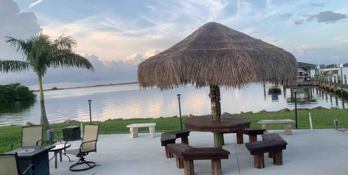 An outdoor space features a thatched tiki table surrounded by several wooden benches. A calm river reflects the sky, with a gentle breeze brushing through nearby palm trees. Soft lighting from lanterns enhances the ambiance along the concrete patio overlooking the water.