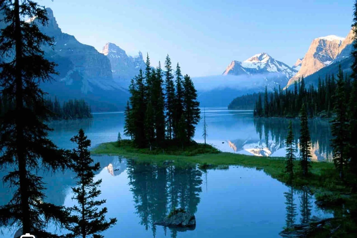 A tranquil lake is surrounded by tall trees and majestic mountains. The water reflects the landscape, creating a mirror-like effect. Soft morning light illuminates the scene, highlighting the natural beauty and serenity of the surrounding wilderness.