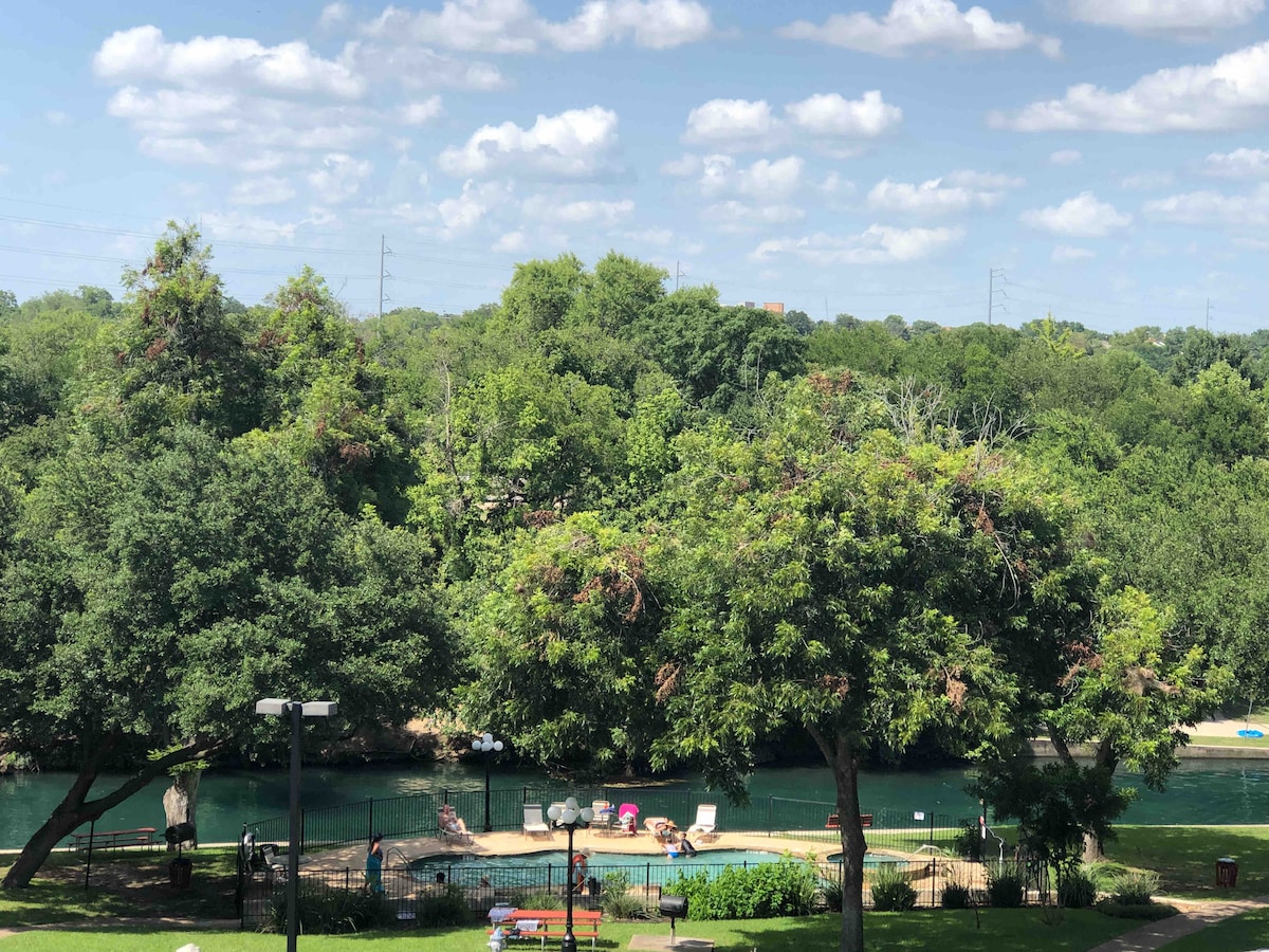 A serene view is presented, showcasing a swimming pool surrounded by lush greenery. The pool area features lounge chairs, while the Comal River flows gently in the background, bordered by mature trees and a clear blue sky dotted with clouds.