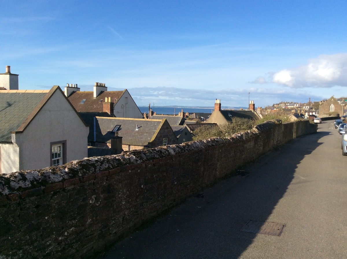 A scenic view of village rooftops extends towards the horizon, where the sea is visible. The image captures a blend of traditional architecture with stone and slate roofs, under a clear blue sky. A quiet road runs alongside a weathered stone wall.
