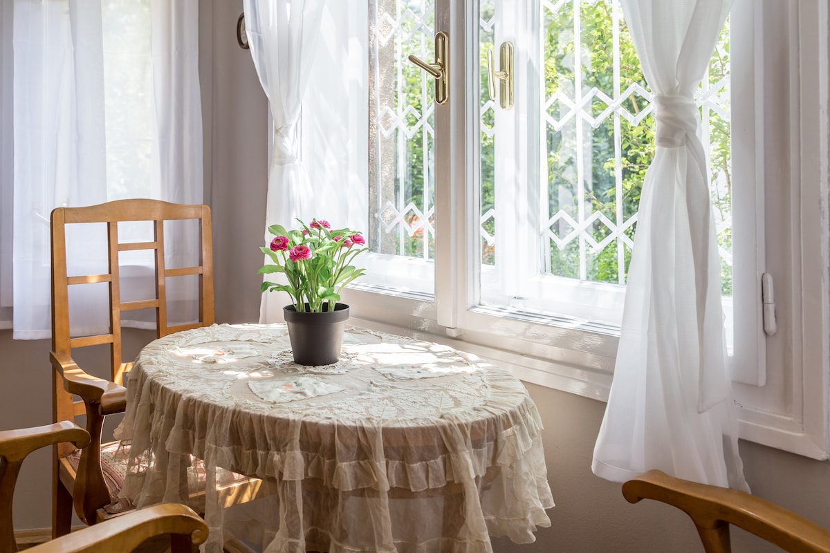 A corner table is positioned by a set of large windows adorned with sheer white curtains, allowing natural light to fill the space. A small potted plant sits at the center of the table, while two wooden chairs are placed beside it.