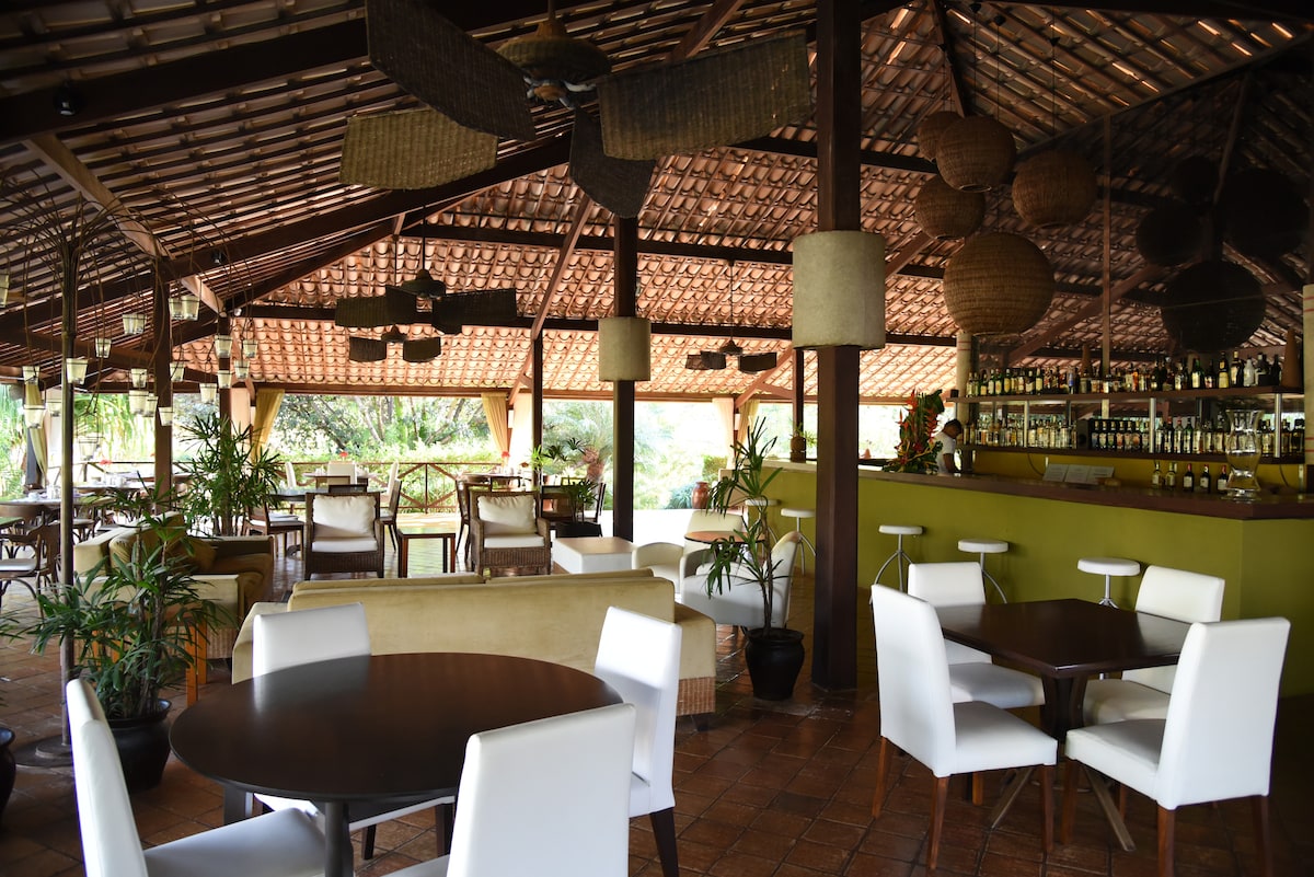 An open dining area features wooden beams and a high ceiling with wicker fans. Tables with dark wooden surfaces are arranged alongside white chairs. A bar with a green accent wall displays various bottles, while plants add a touch of greenery throughout the space.