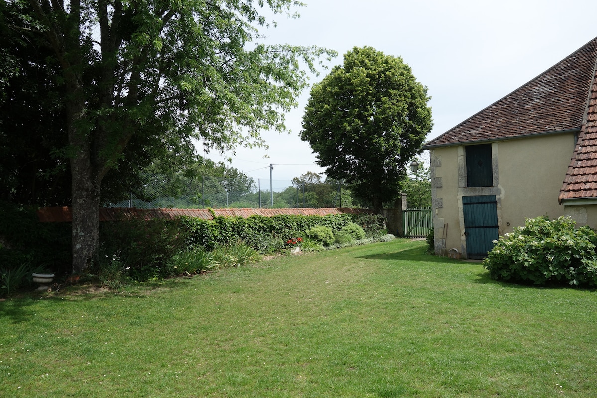 A garden area is presented, featuring well-maintained grass and a mixture of trees and shrubs. A section of the stone building is visible, with a blue door contrasting against the gray facade. The backdrop includes a fence and distant trees, contributing to a serene environment.