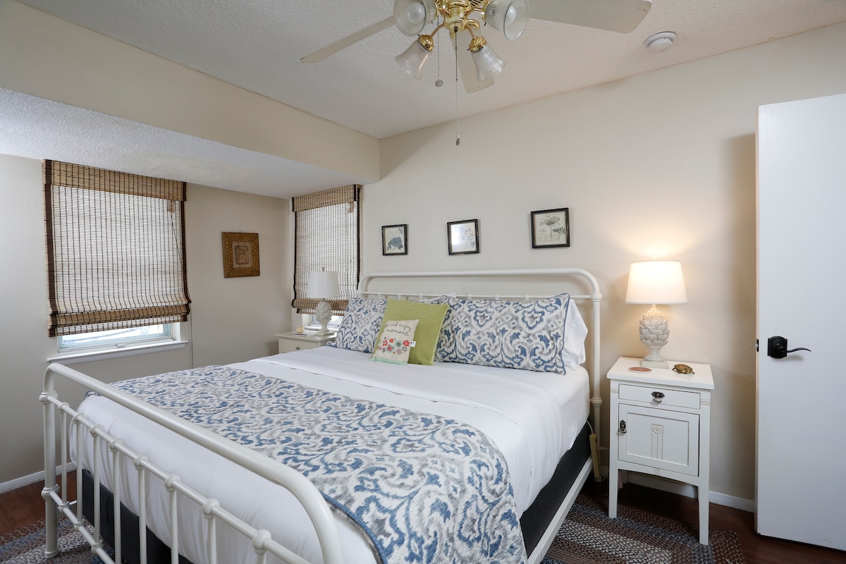 A bright bedroom features a white metal bed frame with a neatly made bed, covered in a blue and white patterned comforter. A bedside table with a lamp sits next to the bed, and natural light enters through partially covered windows with woven shades.