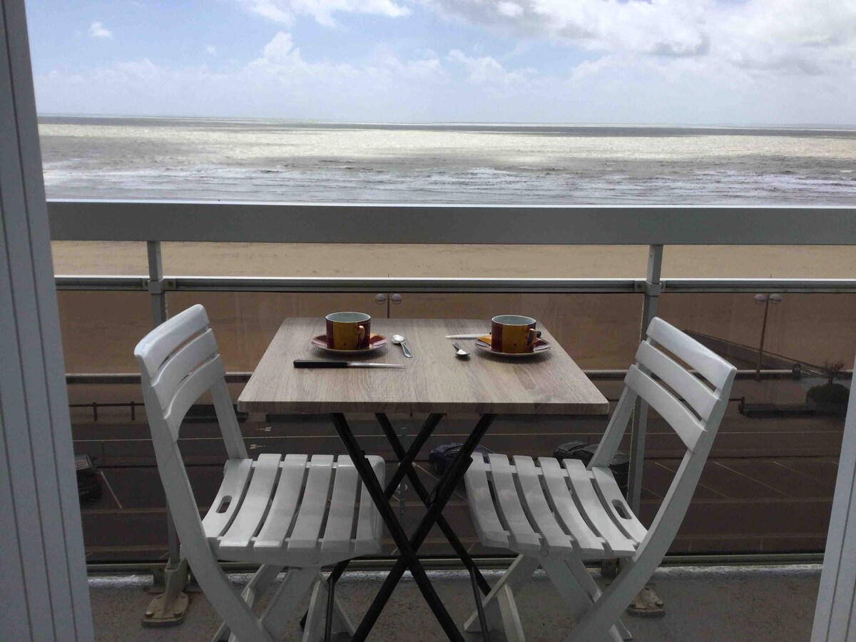 A small balcony features a wooden table set for two, with two cups placed on bright ceramic saucers. Two white chairs are positioned around the table, offering a clear view of the sandy beach and the sea beyond, under a partly cloudy sky.