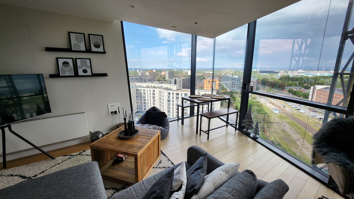 A bright living space features floor-to-ceiling windows, showcasing panoramic views of the city. A comfortable gray couch complements a wooden coffee table, while a small dining table is situated nearby. Modern artwork is displayed on the walls, enhancing the contemporary aesthetic.