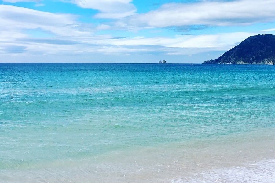 A serene beach scene features soft sand meeting clear, multi-toned blue waters. Gentle waves create a tranquil rhythm as a distant green hill is visible along the coastline under a cloudy sky. The expansive horizon blends seamlessly with the water.
