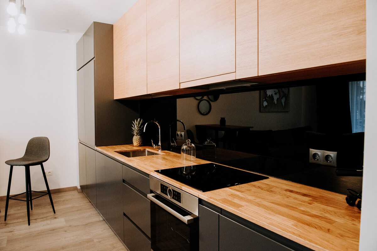 A modern kitchen is showcased with a sleek design featuring dark cabinetry and a light wooden countertop. Essential appliances include a stovetop, sink, and a stylish barstool is positioned nearby, enhancing the functional space.