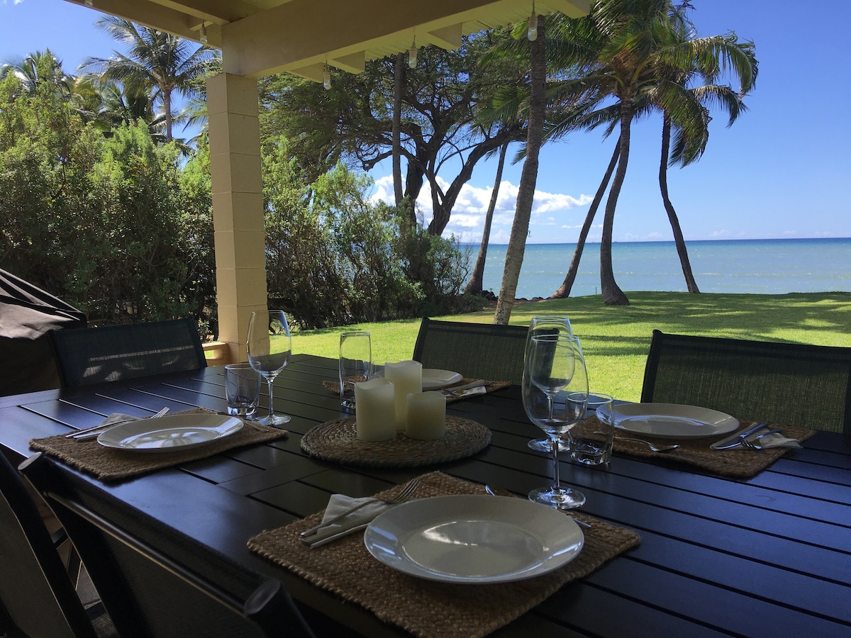 An outdoor dining table is set for four, featuring beige plates and glassware. A centerpiece of simple white candles complements the natural setting. Lush greenery and palm trees frame the view of the ocean in the background, creating a serene ambiance.
