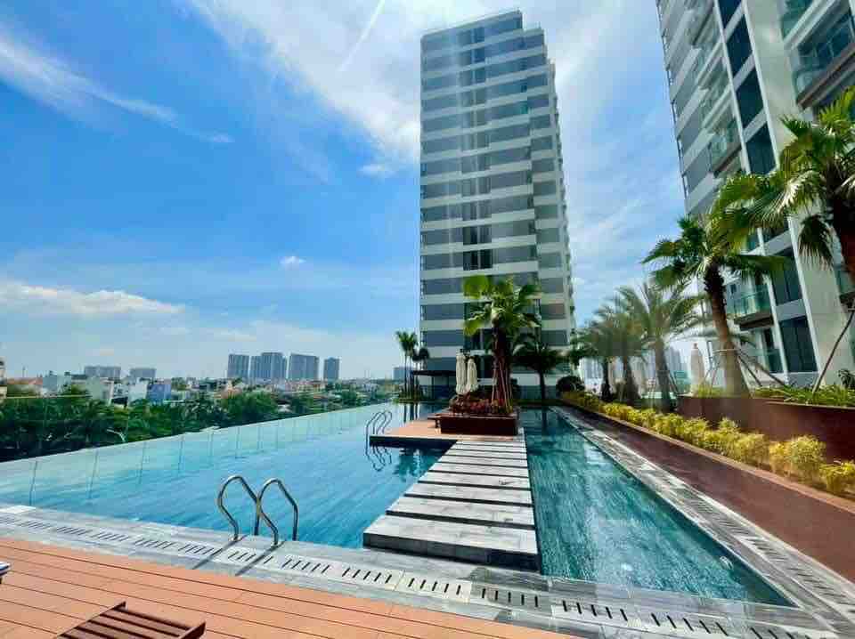 An infinity pool is presented on a rooftop, framed by modern high-rise buildings and lush greenery. Comfortable lounge chairs line the pool area, while palm trees provide a touch of nature against the bright blue sky. The surrounding cityscape is observable in the background.