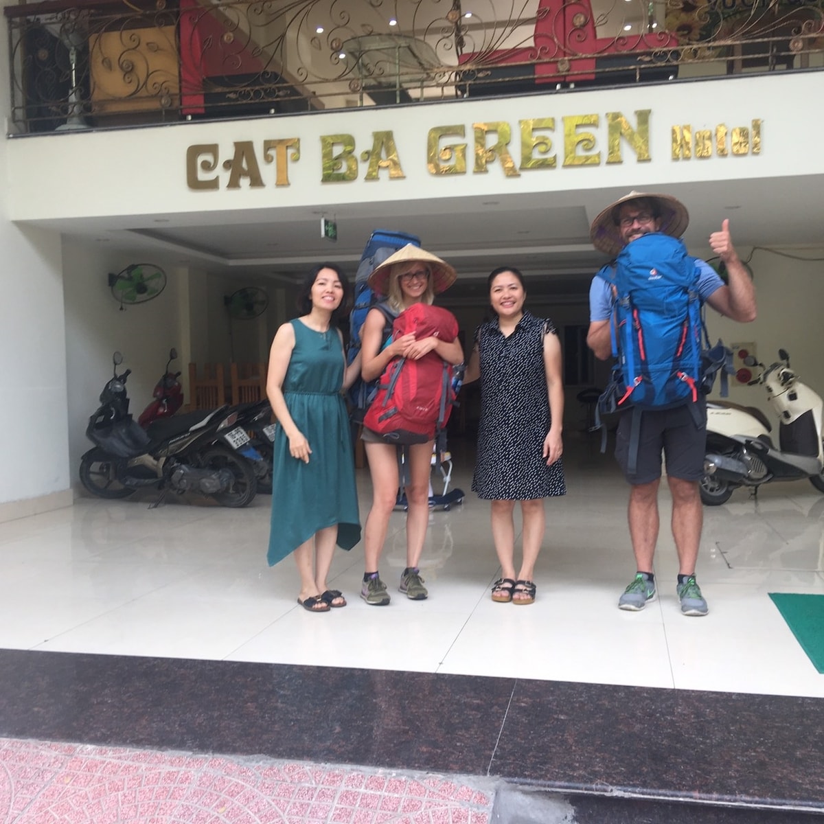 A welcoming entrance to Cat Ba Green Hotel is visible, featuring a spacious lobby area with modern decor. The hotel name is prominently displayed in gold lettering above, and various bicycles and motorcycles can be seen parked nearby.