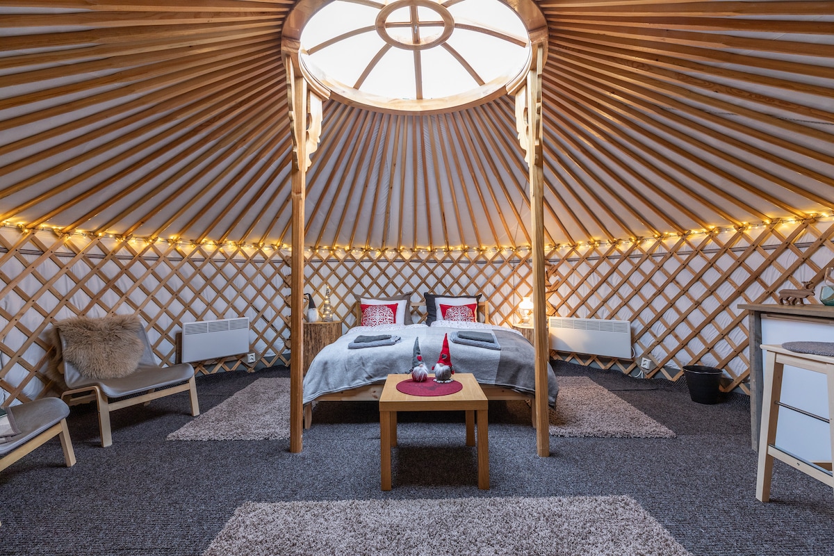 A spacious yurt interior features a circular skylight surrounded by wooden beams. A cozy bed is positioned centrally, accompanied by decorative pillows. Two lounge chairs and a small table enhance the warm atmosphere, with soft carpeting underfoot and gentle lighting accentuating the unique design.
