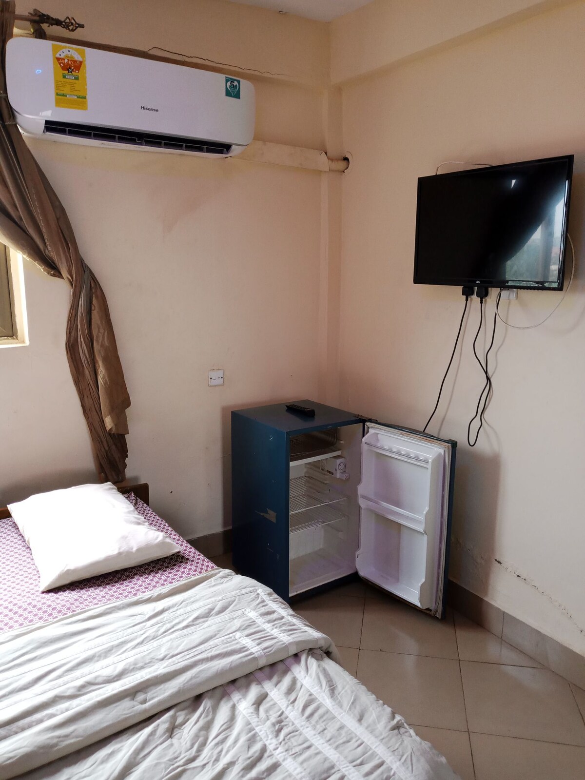 The room features a spacious bed topped with a single pillow and a soft blanket. A small refrigerator is positioned beside the bed, with its door slightly ajar. A flat-screen television is mounted on the wall, and a window provides natural light filtered through partially drawn curtains.