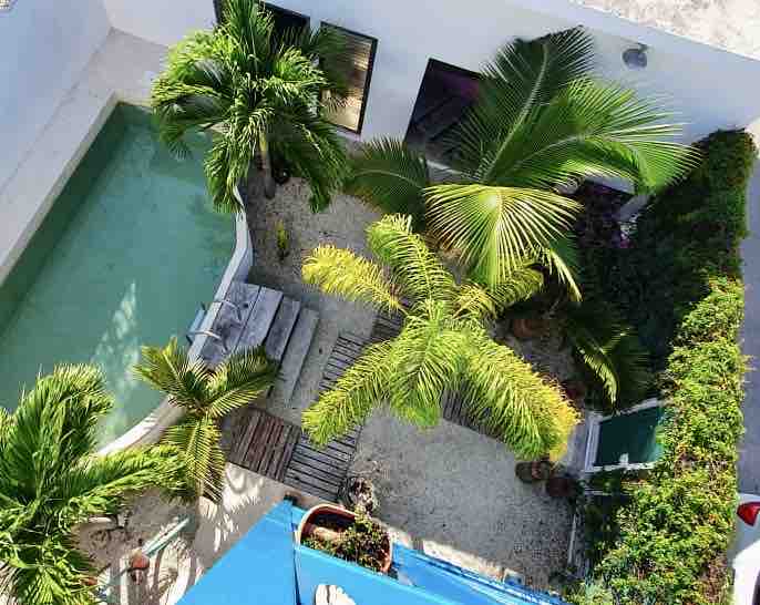 An aerial view of a private garden features a turquoise pool surrounded by palm trees and lush greenery. A wooden deck leads from the pool, and pathways are lined with gravel. Colorful plants add vibrancy to the serene outdoor space.