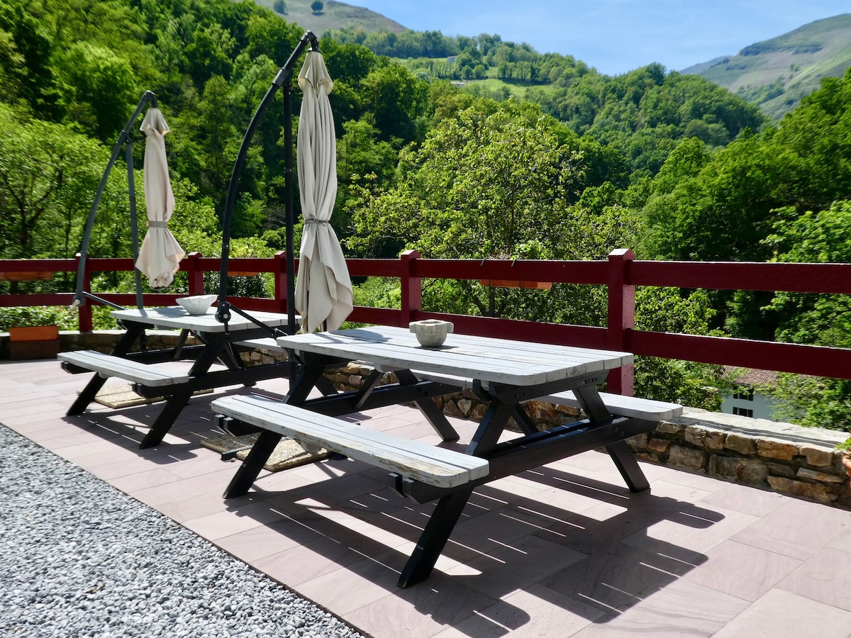 An outdoor terrace features two picnic tables with attached shaded umbrellas, set against a backdrop of lush green hills. The space is paved with smooth stone and surrounded by a red railing, providing a serene environment for relaxation.
