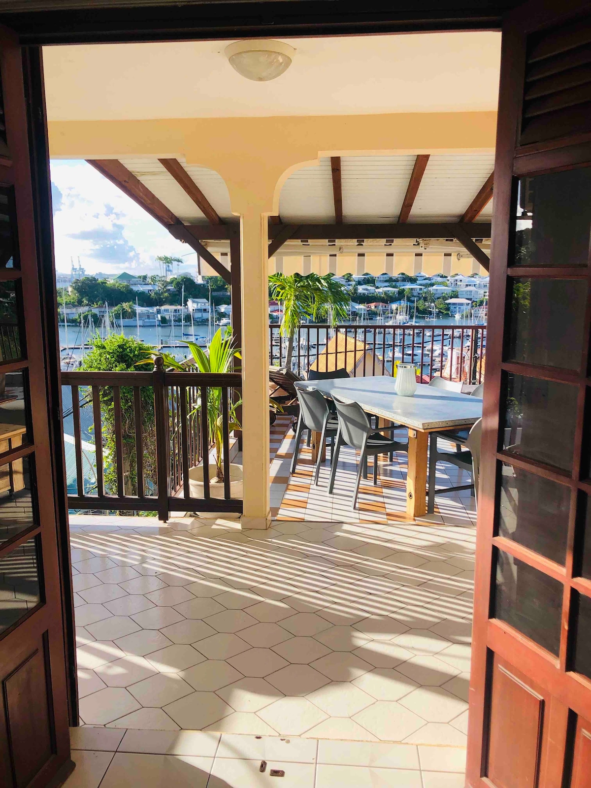 A view from an open doorway reveals a spacious terrace with a wooden dining table and six chairs. Lush greenery is seen alongside the railing, and the marina is visible in the background. Natural light filters through, casting patterns on the tiled floor.