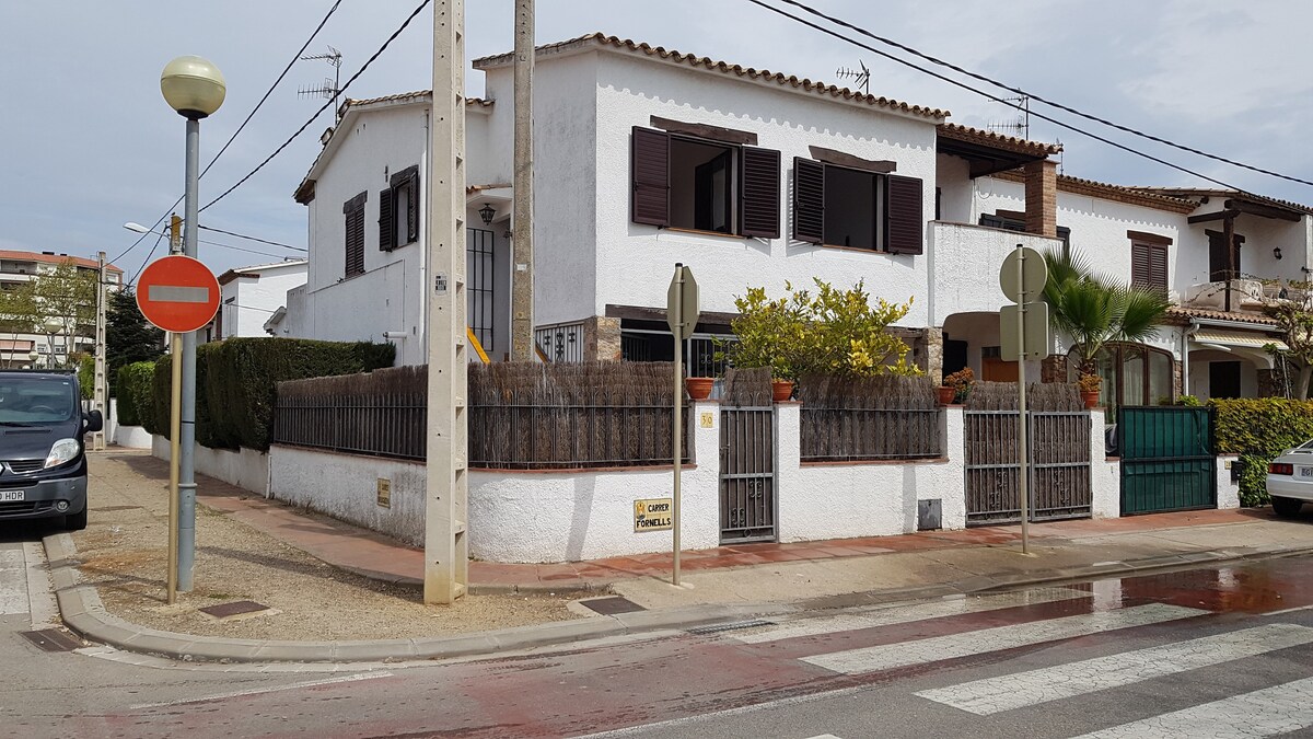 A two-story white building is visible, featuring wooden shutters and a fenced front yard with green plants. The entrance is marked by a set of stairs leading to the doorway. Pedestrian pathways and street signs can be seen in the foreground, indicating the residential area.