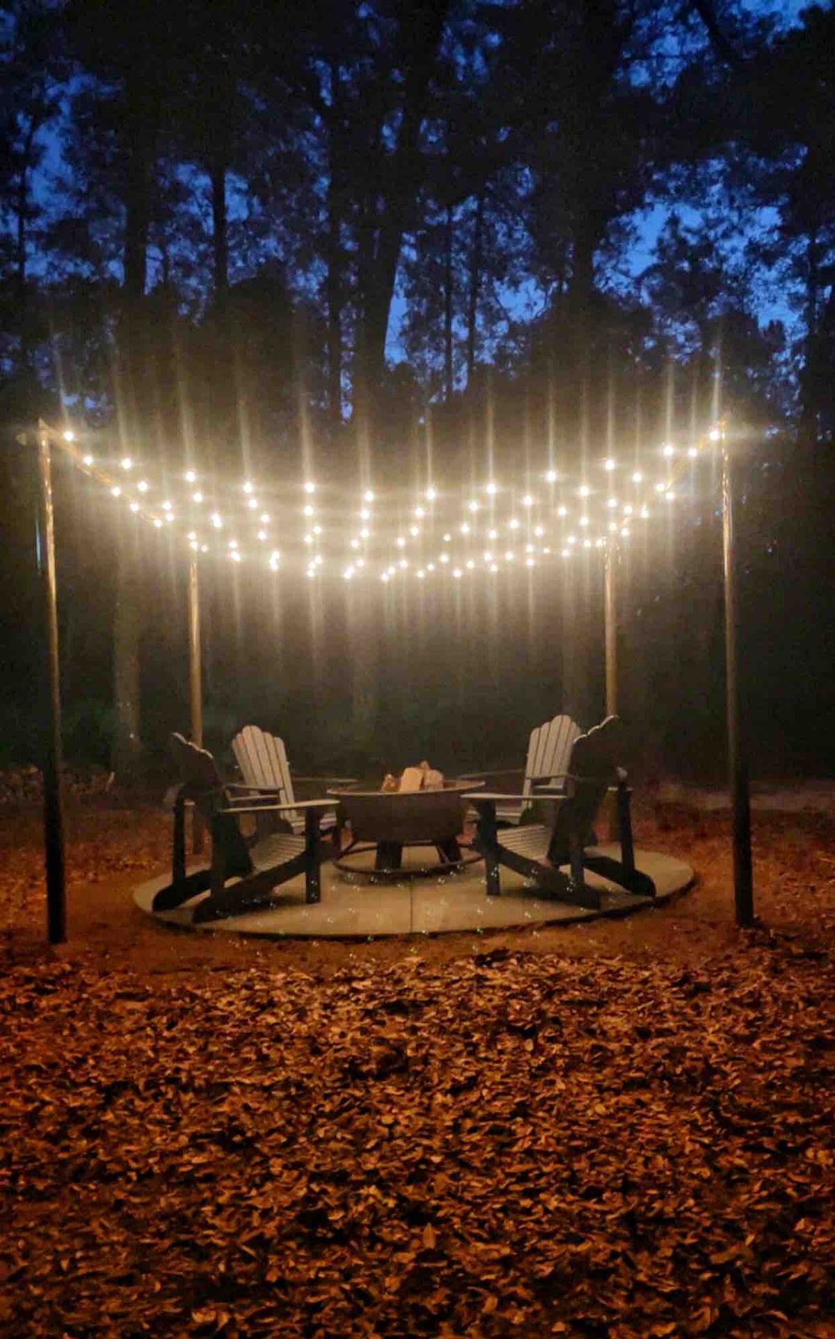 A circular seating area is surrounded by a natural setting, featuring several adirondack chairs arranged around a fire pit. String lights are suspended above, creating a soft glow against the evening sky. The ground is covered in leaves, enhancing the rustic charm.