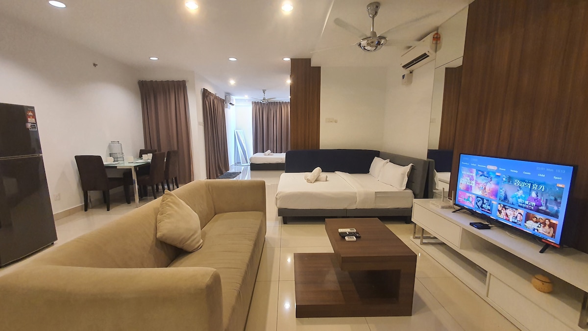 An open living area features a spacious sofa and wooden coffee table, with a large television mounted on an accent wall. A dining table is visible, with a refrigerator in the corner. Natural light enters through floor-to-ceiling curtains, illuminating the comfortable sleeping area.